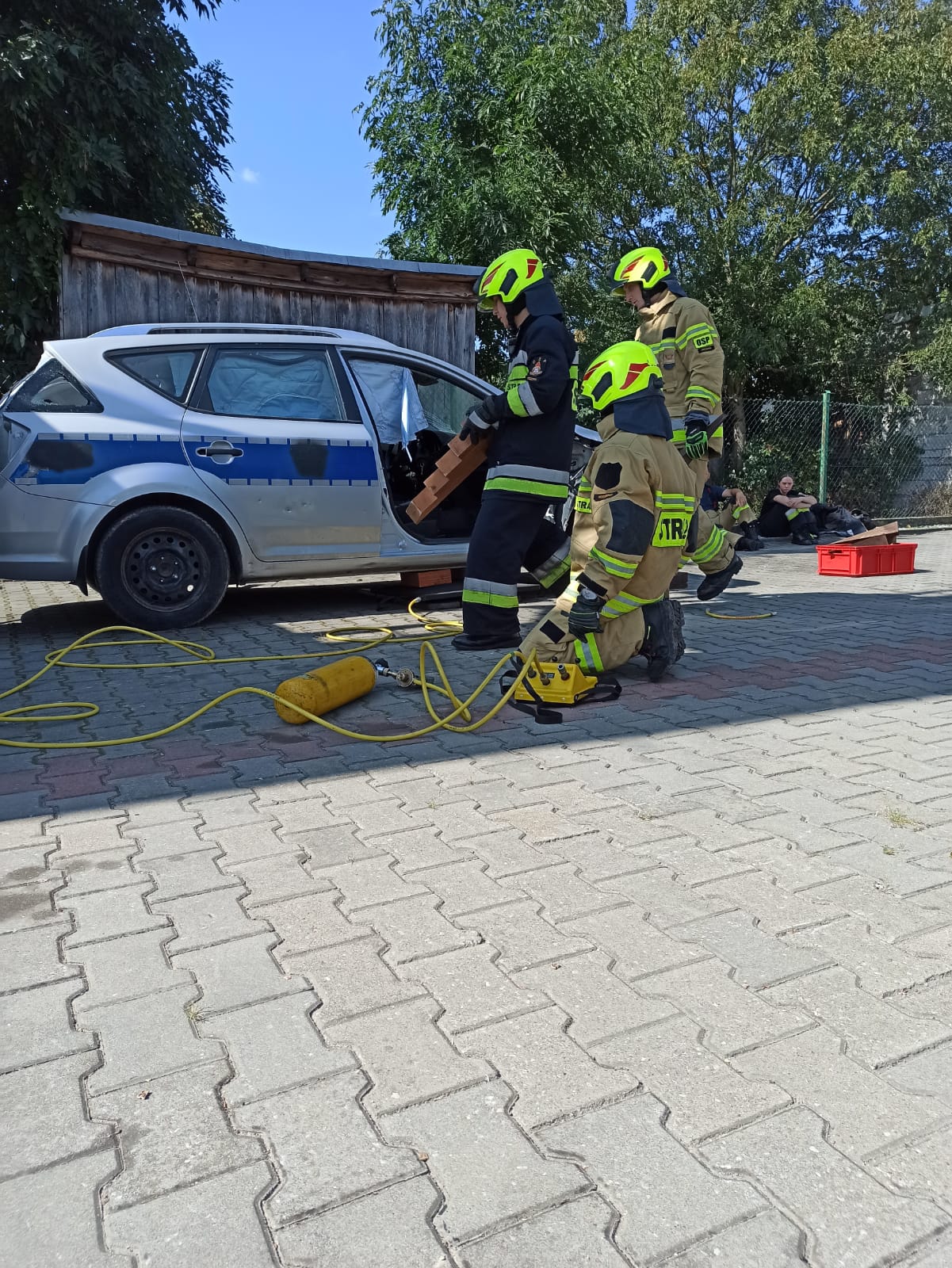 Trzech strażaków ubranych w ubrania specjalne ćwiczą przy wraku samochodu policyjnego używając zestawów pneumatycznych.