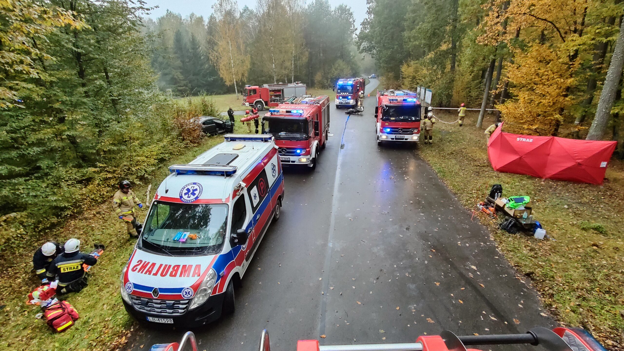 Cztery pojazdy pożarnicze i ambulans ustawione na leśnej drodze. Strażacy podzieleni na grupy ćwiczą udzielanie pierwszej pomocy przedmedycznej. Po lewej stronie w krzakach znajduje się rozbity samochód osobowy. Po prawej stronie rozstawiony jest parawan