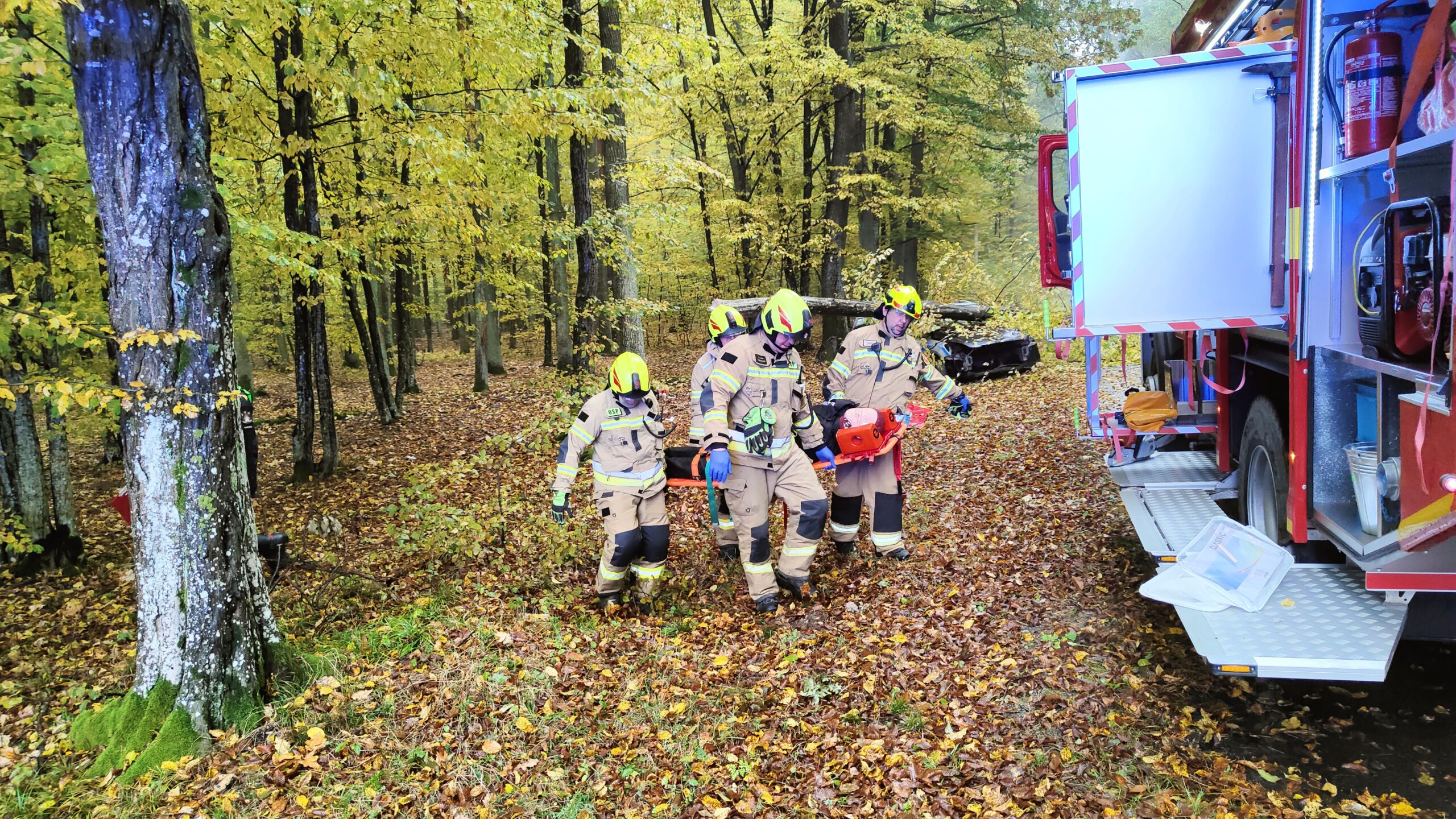 Strażacy niosą manekina ćwiczebnego na noszach typu deska. Z Prawej strony stoi samochód strażacki z pootwieranymi skrytkami sprzętowymi.