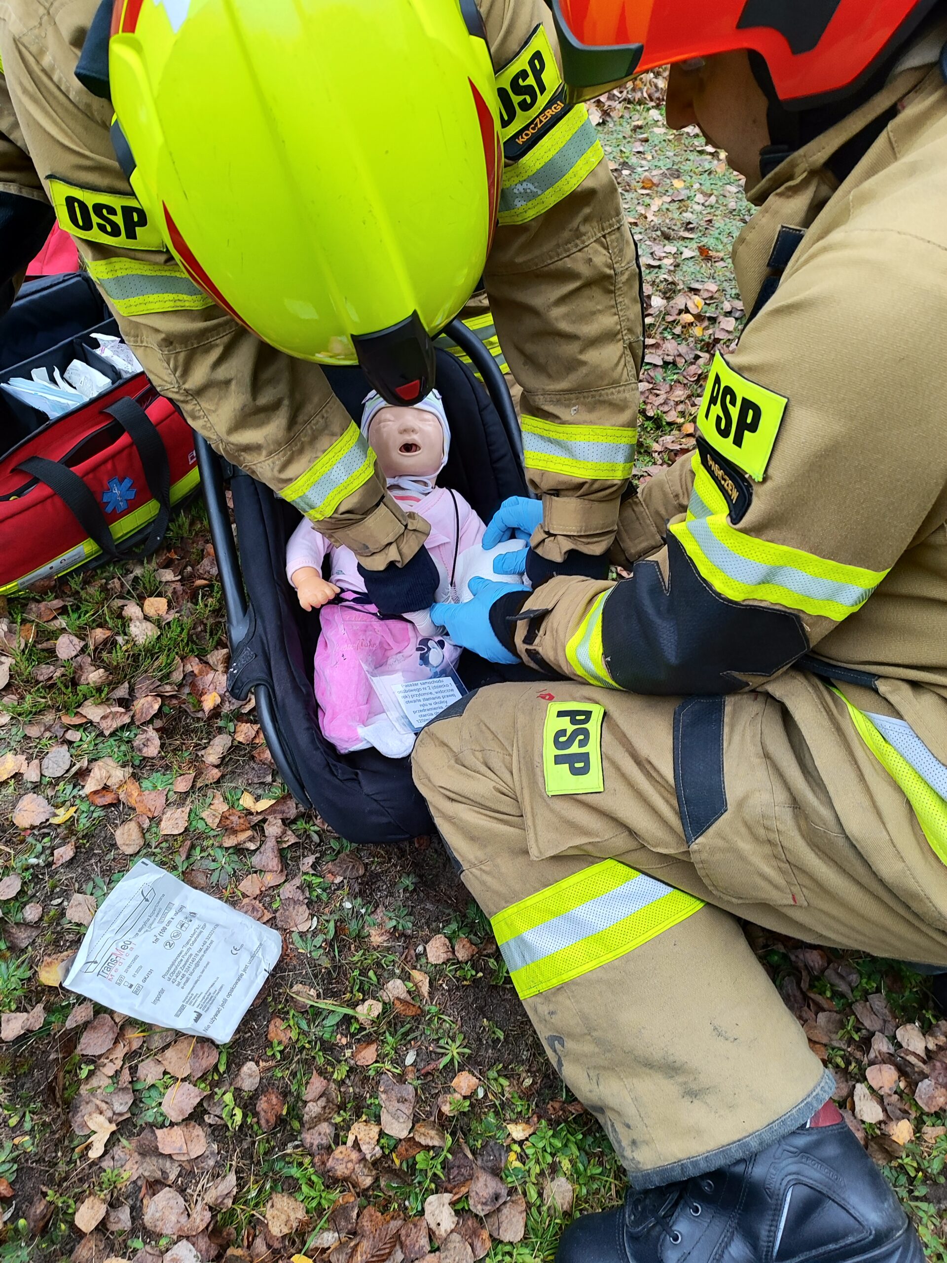 Dwóch strażaków ćwiczy udzielanie pierwszej pomocy przedmedycznej dziecku znajdującemu się w nosidełku