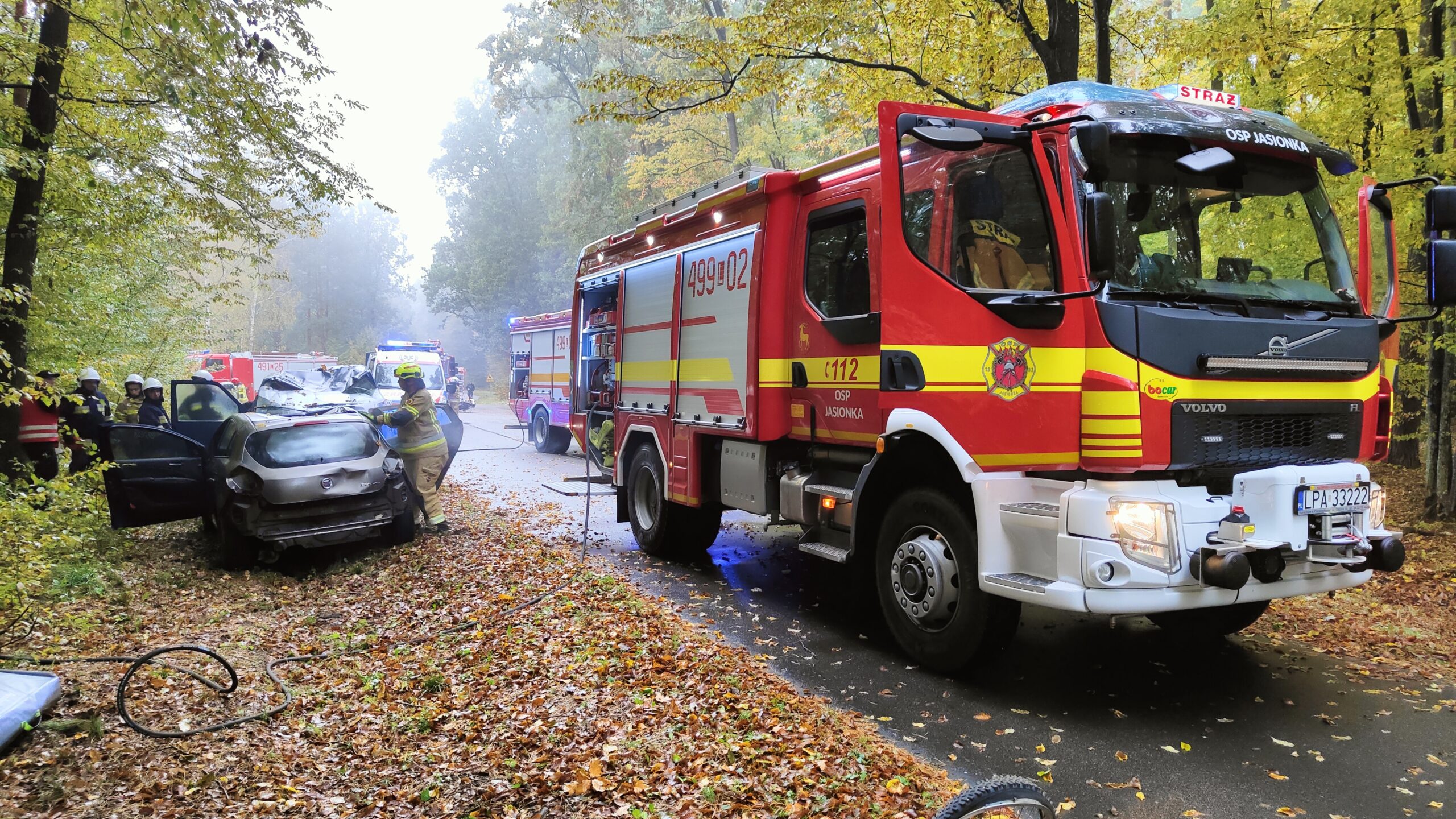 Pojazdy Pożarnicze stoją na leśnej drodze. po lewej stronie znajduje się auto osobowe a strażacy ćwiczą wydobywanie ofiar wypadków komunikacyjnych