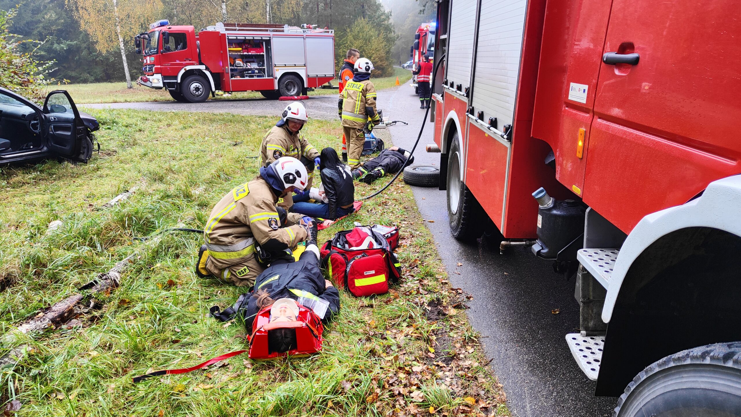 Strażacy ćwiczy udzielanie pierwszej pomocy przedmedycznej. Z prawej strony ustawiony jest samochód pożarniczy. Z tyłu na skrzyżowaniu ustawiony jest kolejny samochód strażacki. Ma otwartą jedną z żaluzji sprzętowych