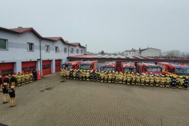 Kilkudziesięciu strażaków stoi w dwuszeregu na placu Komendy Powiatowej PSP w Parczewie. Za nimi ustawione są pojazdy pożarnicze.