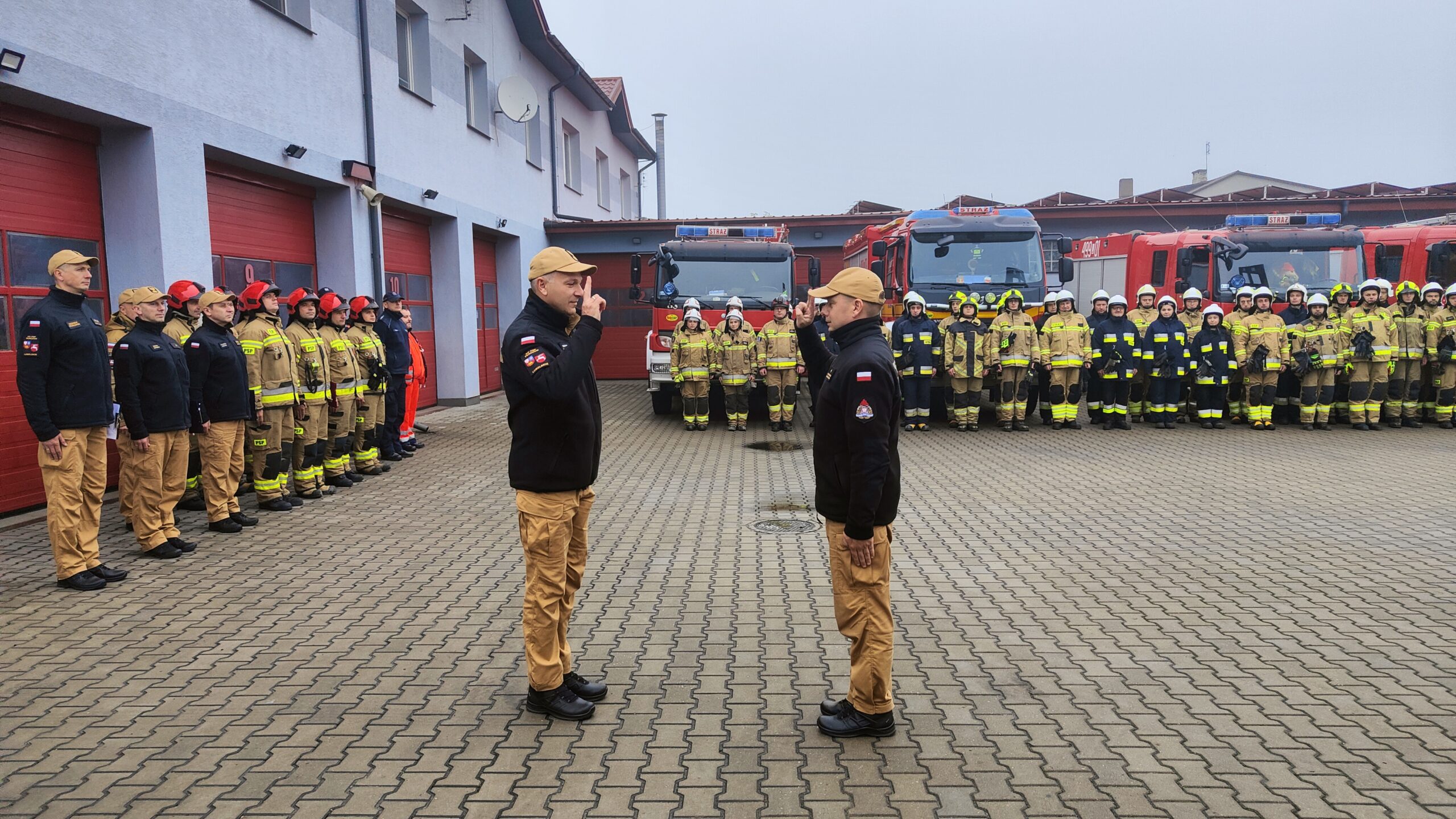 Dwóch strażaków stojących na przeciwko siebie salutują. w oddali strażacy ochotnicy ustawieni w dwuszeregu. Za nimi pojazdy pożarnicze.