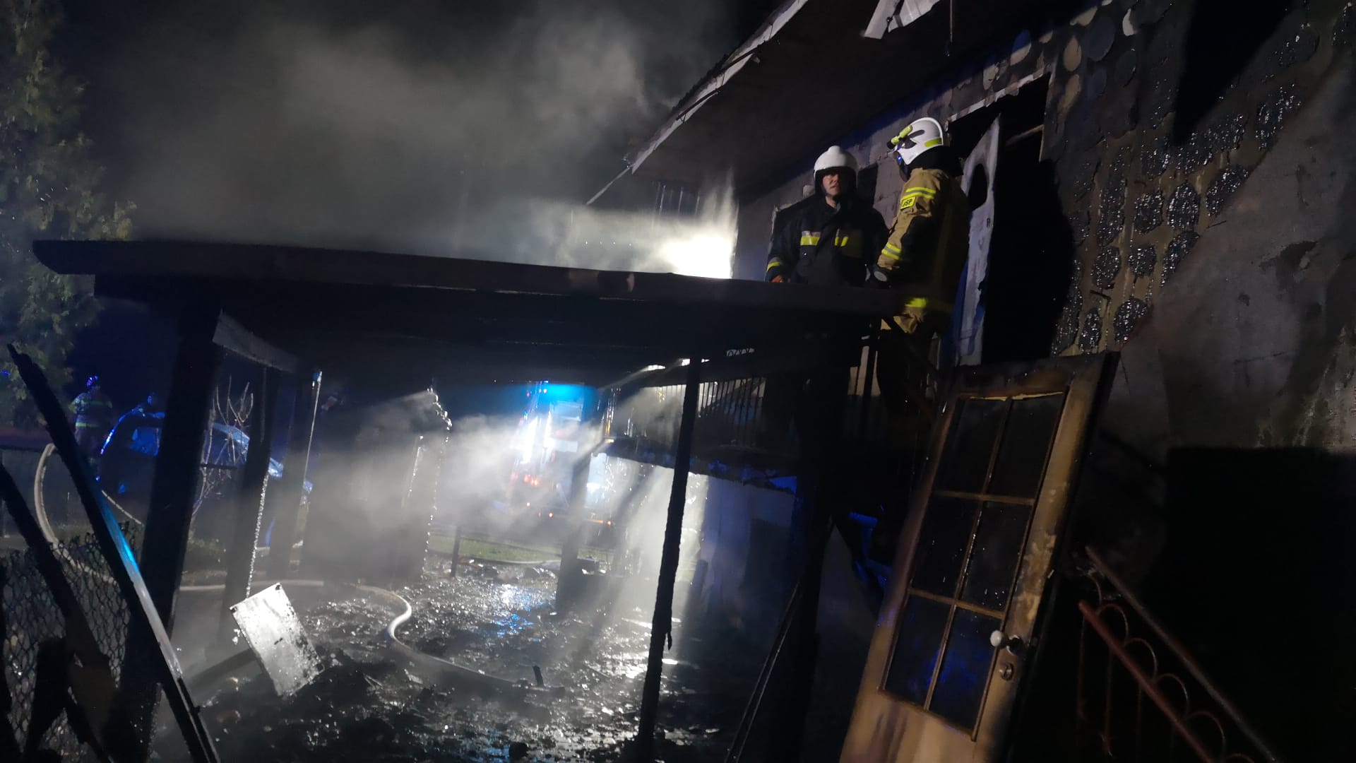 Strażacy stoją przy spalonej części budynku i wiacie.