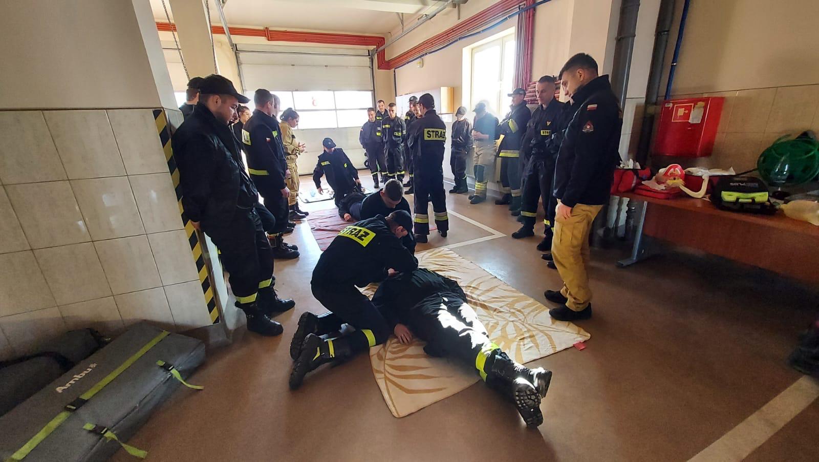 Strażacy uczą się udzielania pierwszej pomocy przedmedycznej. Dwie osoby leży na podłodze a cztery kolejne ćwiczy udzielanie pomocy. Pozostali strażacy obserwują ćwiczenia.