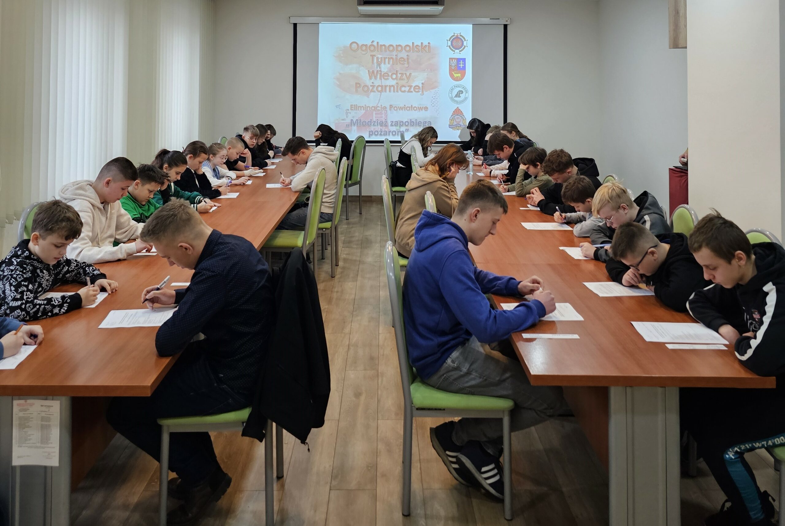 Młodzież siedzi przy długich stołach i rozwiązuje test. Na ścianie w głębi sali wyświetlony jest napis Ogólnopolski Turniej Wiedzy Pożarniczej.