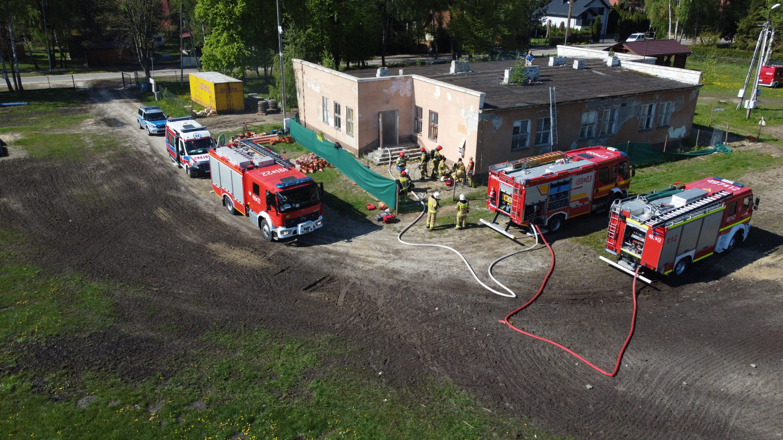 Pozorowany pożar budynku. W ćwiczeniu biorą udział trzy pojazdy pożarnicze, Zespół Ratownictwa Medycznego i Policja. Strażacy rozwinęli linie gaśnicze prowadzące do wnętrza budynku.