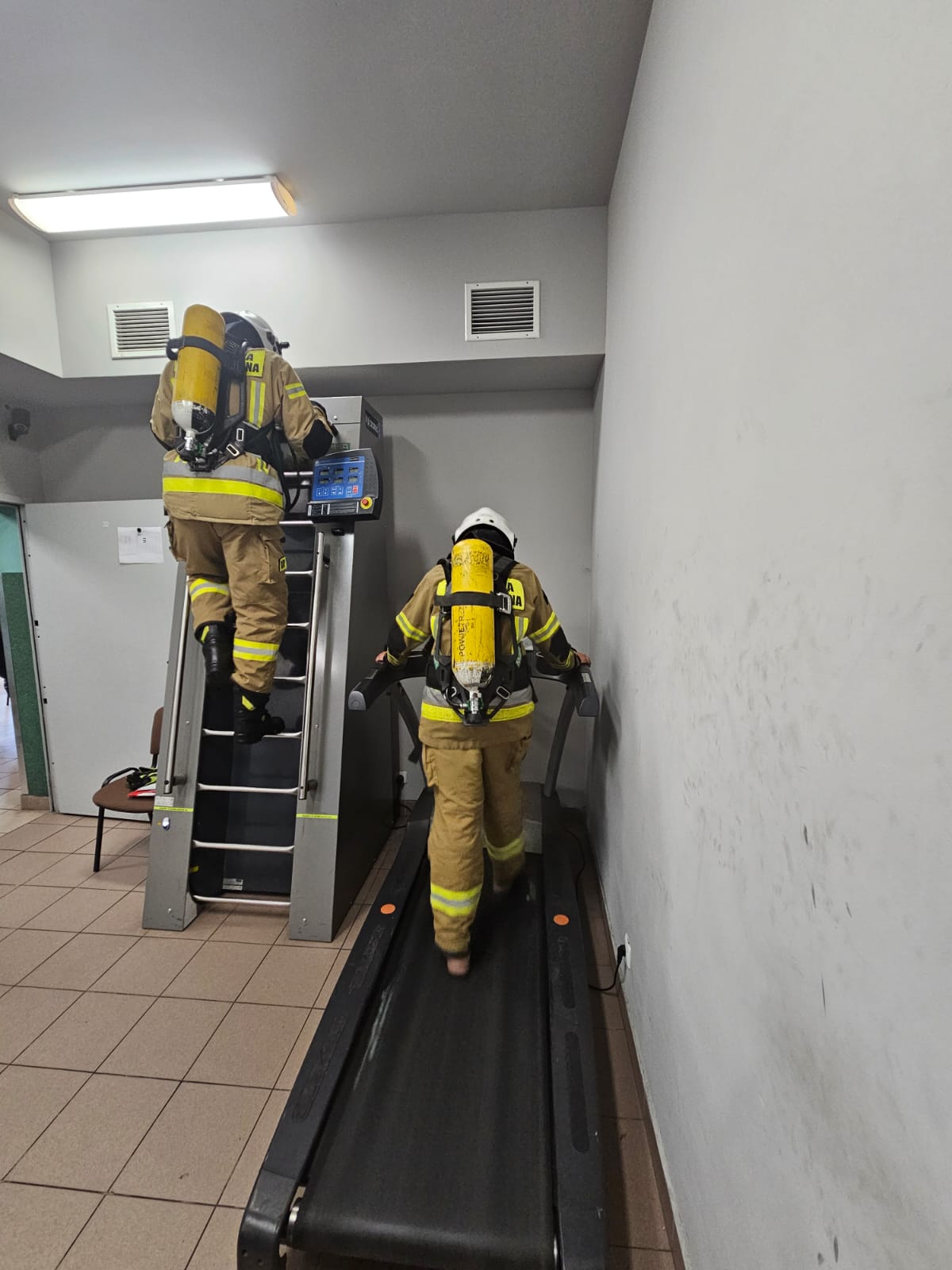 Dwóch strażaków ubranych w ubrania specjalne i aparaty ochrony dróg oddechowych wykonują ćwiczenia na ścieżce wydolnościowej w komorze dymowej