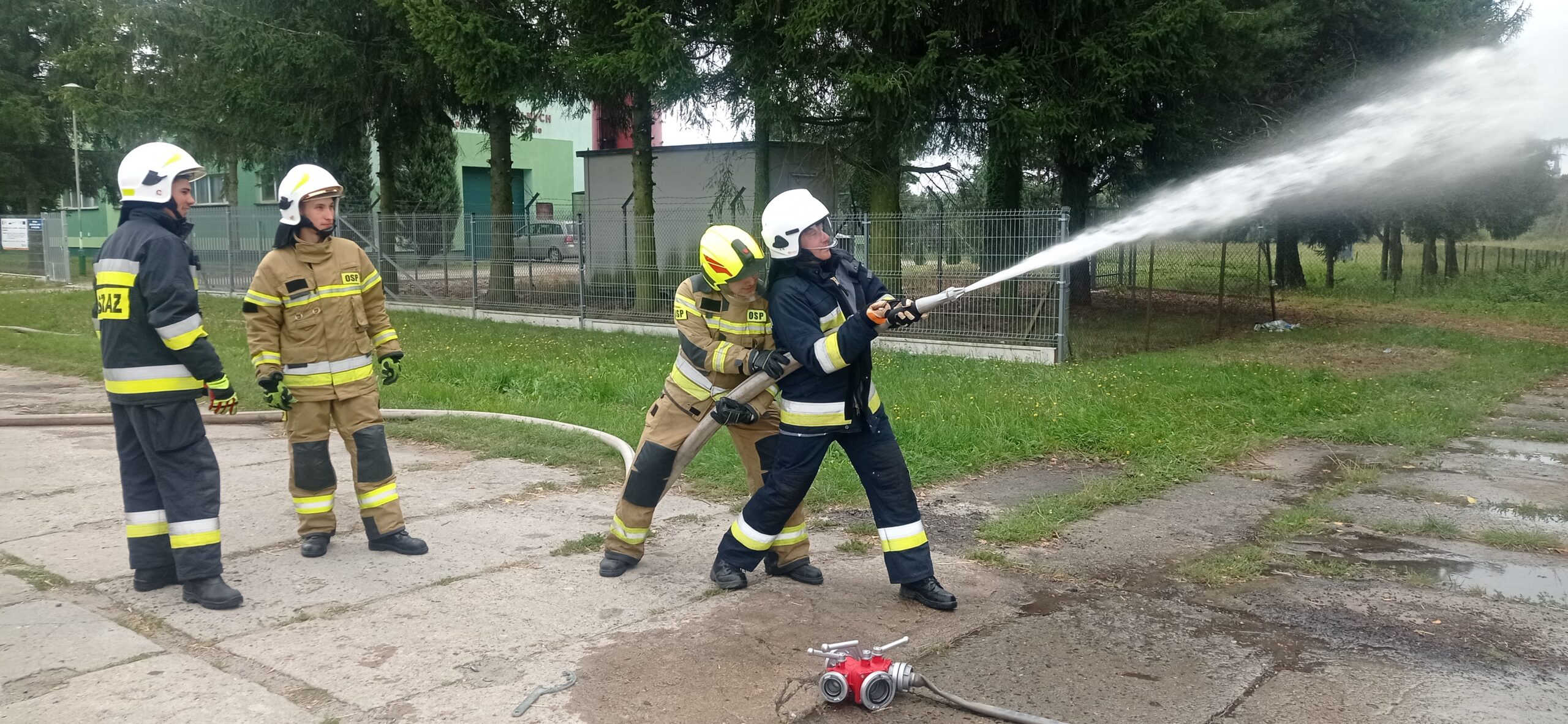 Dwóch strażaków ćwiczy podawanie prądów gaśniczych wody. Dwóch kolejnych obserwuje to Ćwiczenie.