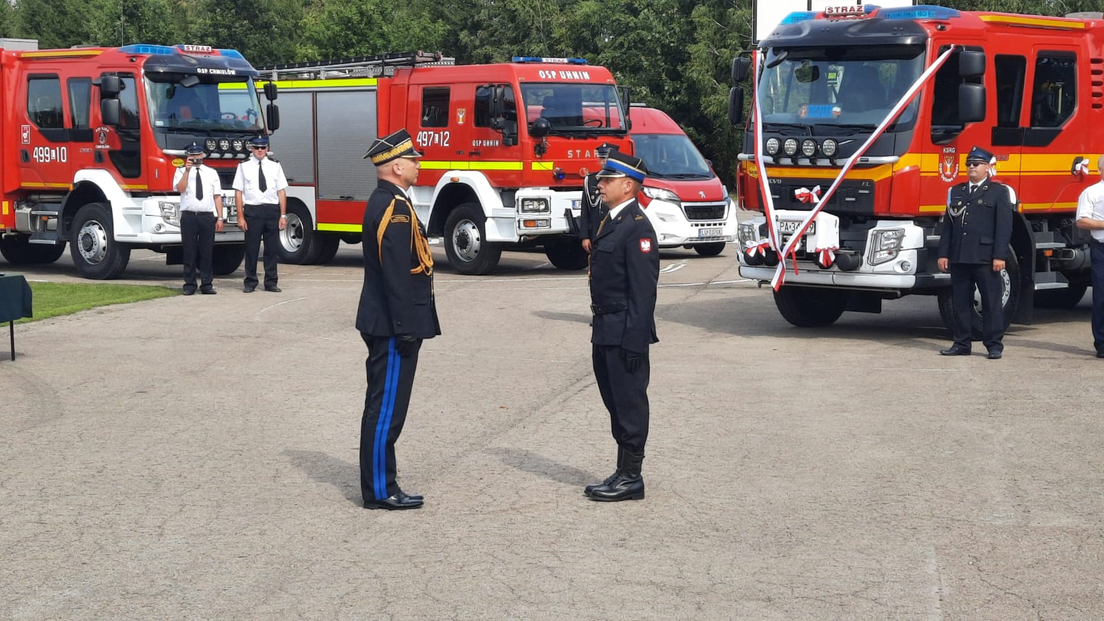 Dwóch strażaków ubranych w mundury wyjściowe stoi na przeciwko siebie. Na drugim planie cztery samochody strażackie, jeden z nich z przodu przewieszony jest biało-czerwoną wstęga. obok samochodów stoi trzech strażaków.
