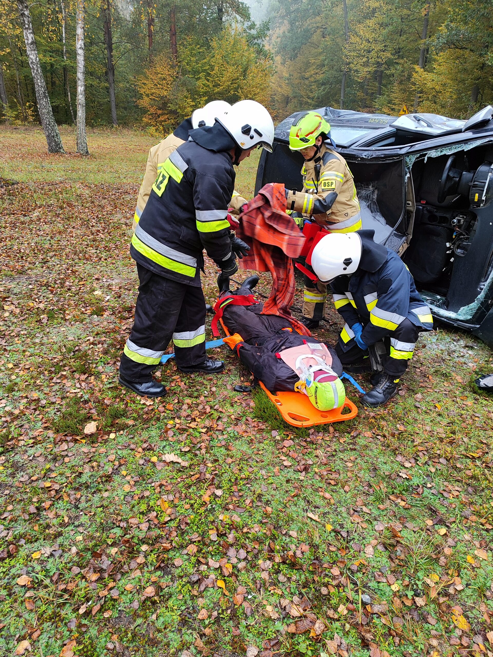 Czterech strażaków ochotników ćwiczą udzielanie pierwszej pomocy przedmedycznej. na ziemi leży deska ratownicza a na niej fantom szkoleniowy. Za strażakami na boku leży rozbity samochód osobowy.