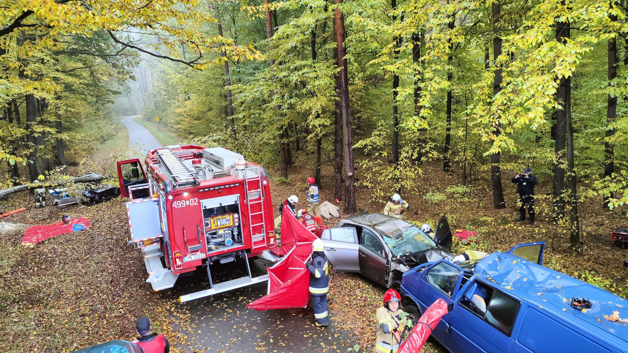 Samochód pożarniczy ustawiony przy wrakach dwóch pojazdów osobowych. Strażacy rozstawiają parawan.