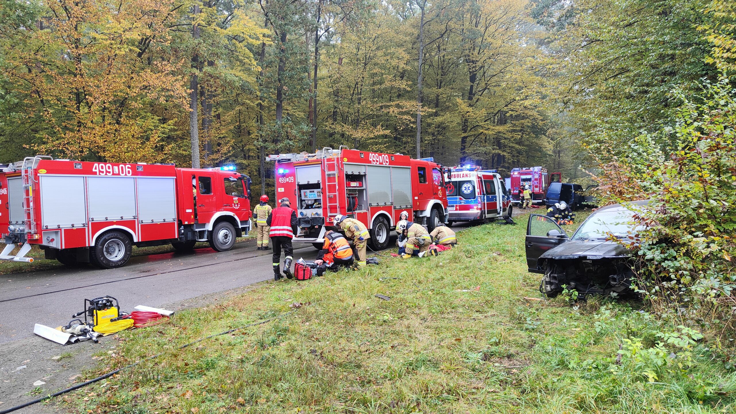Trzy pojazdy pożarnicze i ambulans ustawione na leśnej drodze. strażacy podzieleni na grupy ćwiczą udzielanie pierwszej pomocy przedmedycznej. Po prawej stronie w krzakach znajduje się rozbity samochód osobowy.