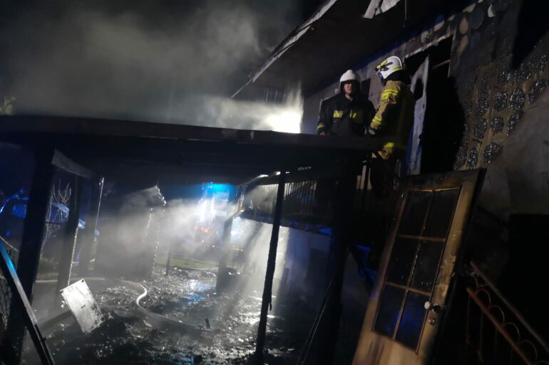 Strażacy stoją przy spalonej części budynku i wiacie.