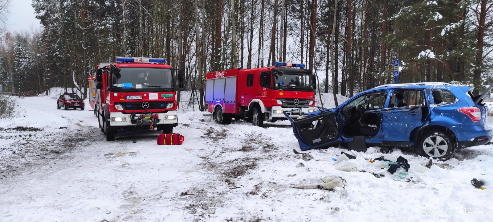 Okres zimowy - spadł śnieg. Uszkodzony samochód osobowy stoi na leśnej drodze. Za nim dwa samochody bojowe straży pożarnej.