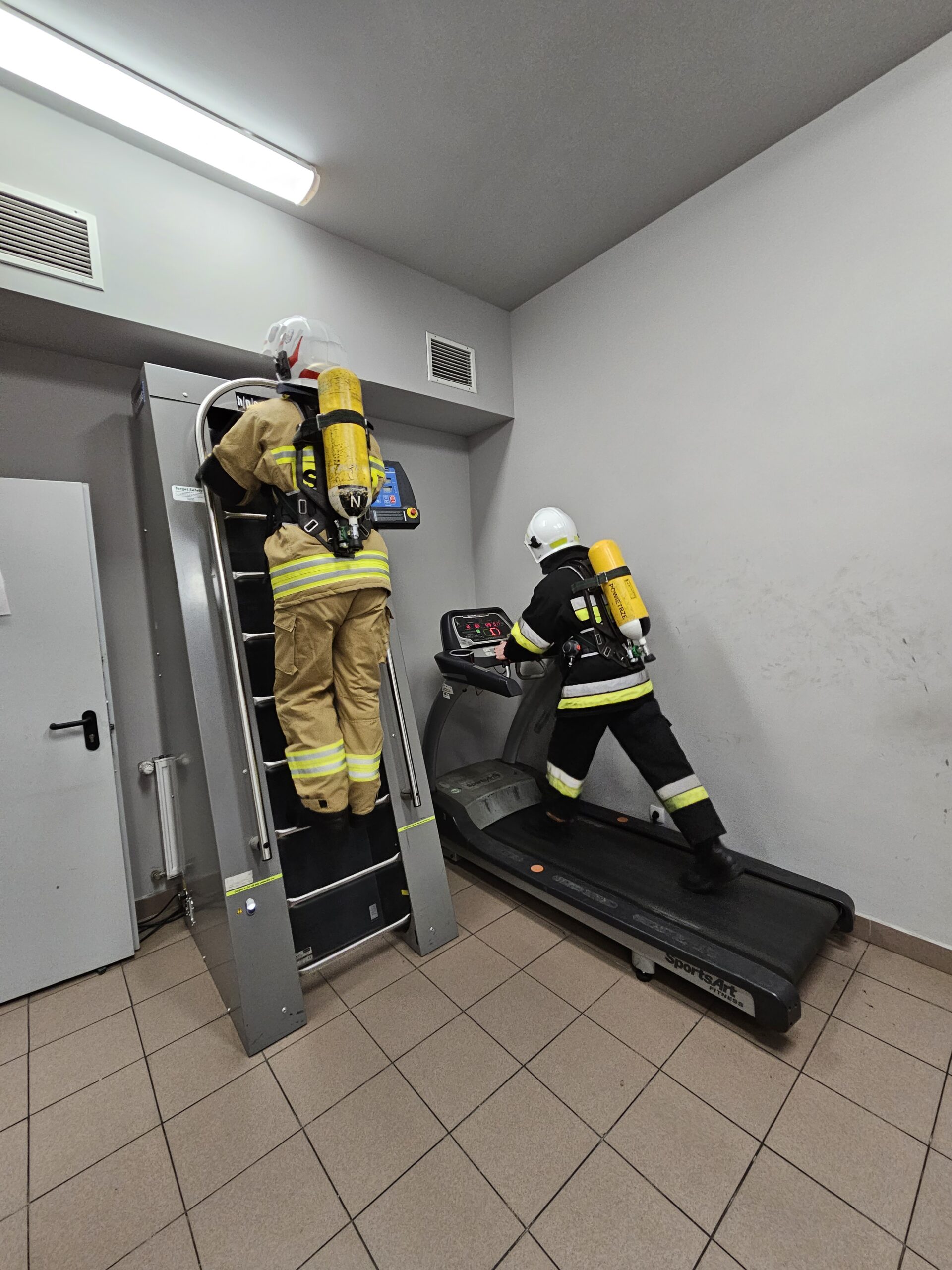 Dwóch strażaków wyposażonych w ubranie specjalne, hełm strażacki i aparat powietrzny ćwiczy swoją wydolność na bieżni i drabinie.