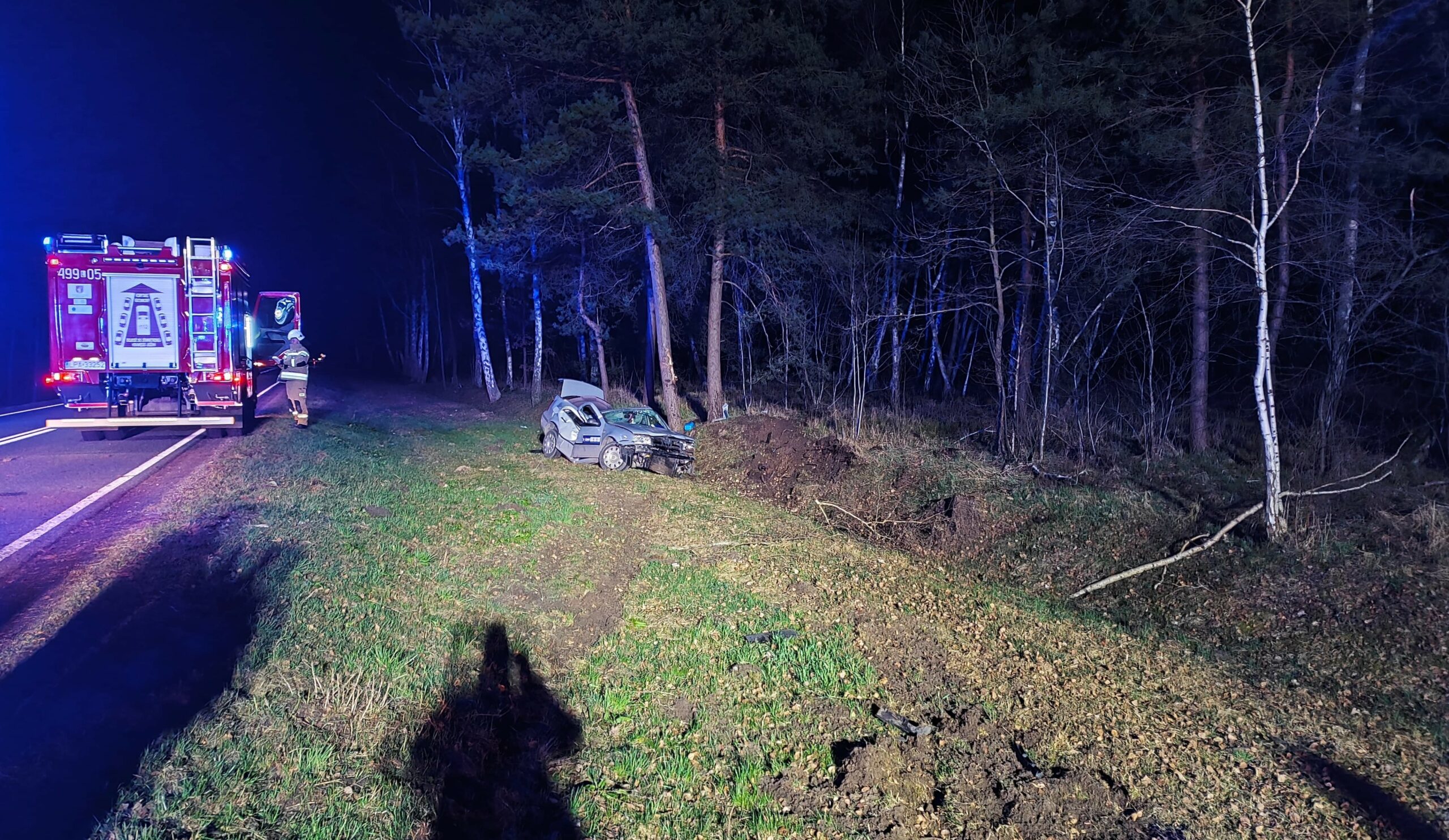 Rozbity samochód osobowy stoi przy pasie drzew poza jezdnią. Z lewej strony stoi samochód pożarniczy a obok niego jeden strażak. Zdarzenie ma miejsce w nocy.