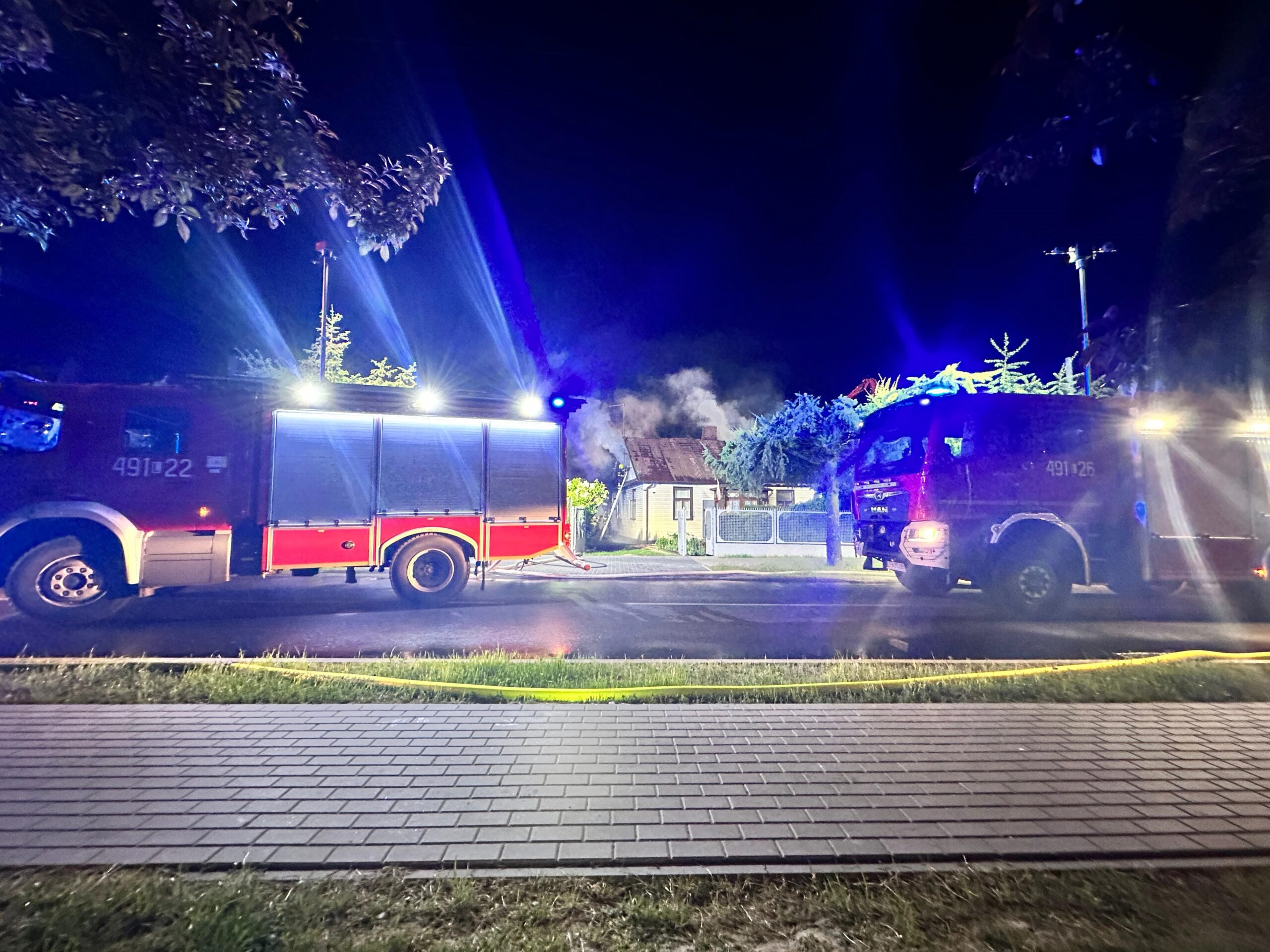 Pora nocna. Dwa pojazdy pożarnicze stoi na jezdni z włączoną sygnalizacją świetlną. w oddali budynek z którego wydobywa się dym.